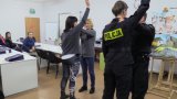 policjanci prezentują elementy samoobrony w trakcie warsztatów w sali przy ul Wolności 101, w tle uczestniczki warsztatów siedzące za stołami