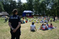 Finał programu ,,Bezpieczne życie” w skansenie, na pierwszym planie widoczna umundurowana policjantka, w tle widoczne są dzieci siedzące na trawie i oglądające spektakl.