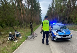 zatrzymani mężczyźni siedzący na poboczu drogi, przed nimi widoczne są rowery oraz funkcjonariusz policji i Straży Granicznej, na jezdni stoi oznakowany radiowóz