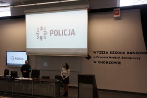 Zdjęcie przedstawia policjanta prowadzącego prelekcję oraz tłumacza językowego.