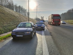 Zdjęcie przedstawia samochód chcącego uniknąć kontroli mężczyzny. W tle policyjny radiowóz oraz przejeżdżające obok inne samochody.