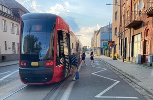 Zdjęcie przedstawia widok z samochodu, który zatrzymał się przed przystankiem, aby umożliwić pieszym dostęp do tramwaju.