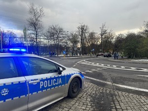 Zdjęcie przedstawia radiowóz, w tle policjanci kierują ruchem na przejściu dla pieszych