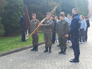 Zdjęcie na zewnątrz szkoły. Przedstawia uczniów ze sztandarem i policjantów.