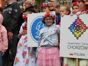 Na zdjęciu grupa dzieci w kolorowych ubraniach. Dziewczynka z wiankiem z kwiatów na głowie. Dwa transparenty. jeden z niebieskim kołem na białym tle. napis niewidoczny drugi z napisem : &quot;Zespół Pieśni i Tańca Chorzów Polska&quot; Źródło: naszemiasto.pl