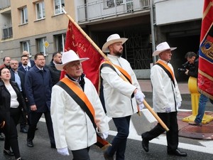 Na zdjęciu trzech mężczyzn idących ze sztandarem . W białych kapeluszach, białych marynarkach, czarnych spodniach. Z pomarańczowo-czarnymi szarfami . Za nimi uczestnicy pochodu. Źródło: naszemiasto.pl