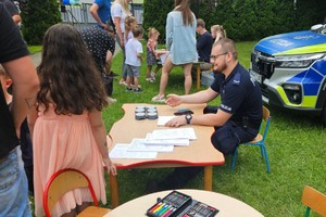 Na zdjęciu siedzący przy stoliku policjant, dzieci i radiowóz