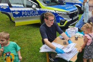 Na zdjęciu mężczyzna siedzący przy stoliku z kartkami na których odbijane są palce na tle radiowozu