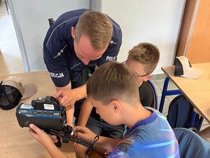 Na zdjęciu dziecko siedzace w ławce trzymajace w ręce miernik prędkości pojazdów i policjant, który tłumaczy dziecku jak działa to urzadzenie