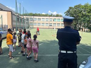Zdjęcie przedstawia policjanta, grupę dzieci ustawionych tyłem oraz jedno dziecko jeżdżące na rowerze po boisku.