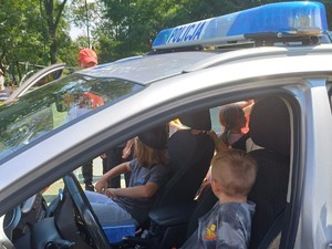 Na zdjęciu dwoje dzieci na przednich siedzeniach radiowozu oraz kilkoro dzieci stojących za radiowozem