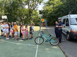 Na zdjęciu grupa dzieci na boisku szkolnym stojąca przed rowerem, policjantką i policjantem oraz samochodem