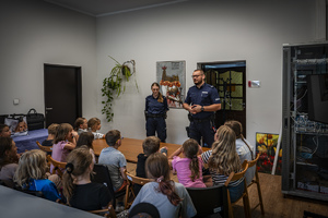 Na zdjęciu policjanta z policjantem stojący przed grupą siedzących przy stole dzieci
