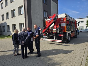 Na zdjęciu Komendant i Zastępca Komendanta Miejskiego Policji w Chorzowie w towarzystwie kobiety i mężczyzny. Za nimi wóz strażacki i maskotka Policji Sznupek