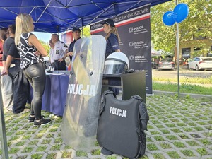 Na zdjęciu dwie policjantki stojące za stołem z materiałami promocyjnymi Policji. O stół oparta jest tarcza z napisem policja oraz kamizelka taktyczna. Przed stołem stoi grupa ludzi
