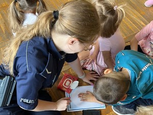 Na zdjęciu dzieci i policjantka siedzące na ziemi. Jedno z dzieci odbija dłoń na kartce papieru