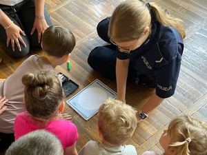 Na zdjęciu dzieci i policjantka siedzące na ziemi. Jedno z dzieci odbija dłoń na kartce papieru