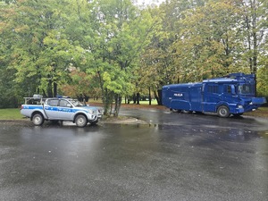 Na zdjęciu radiowóz i armatka wodna stojące pod drzewami.